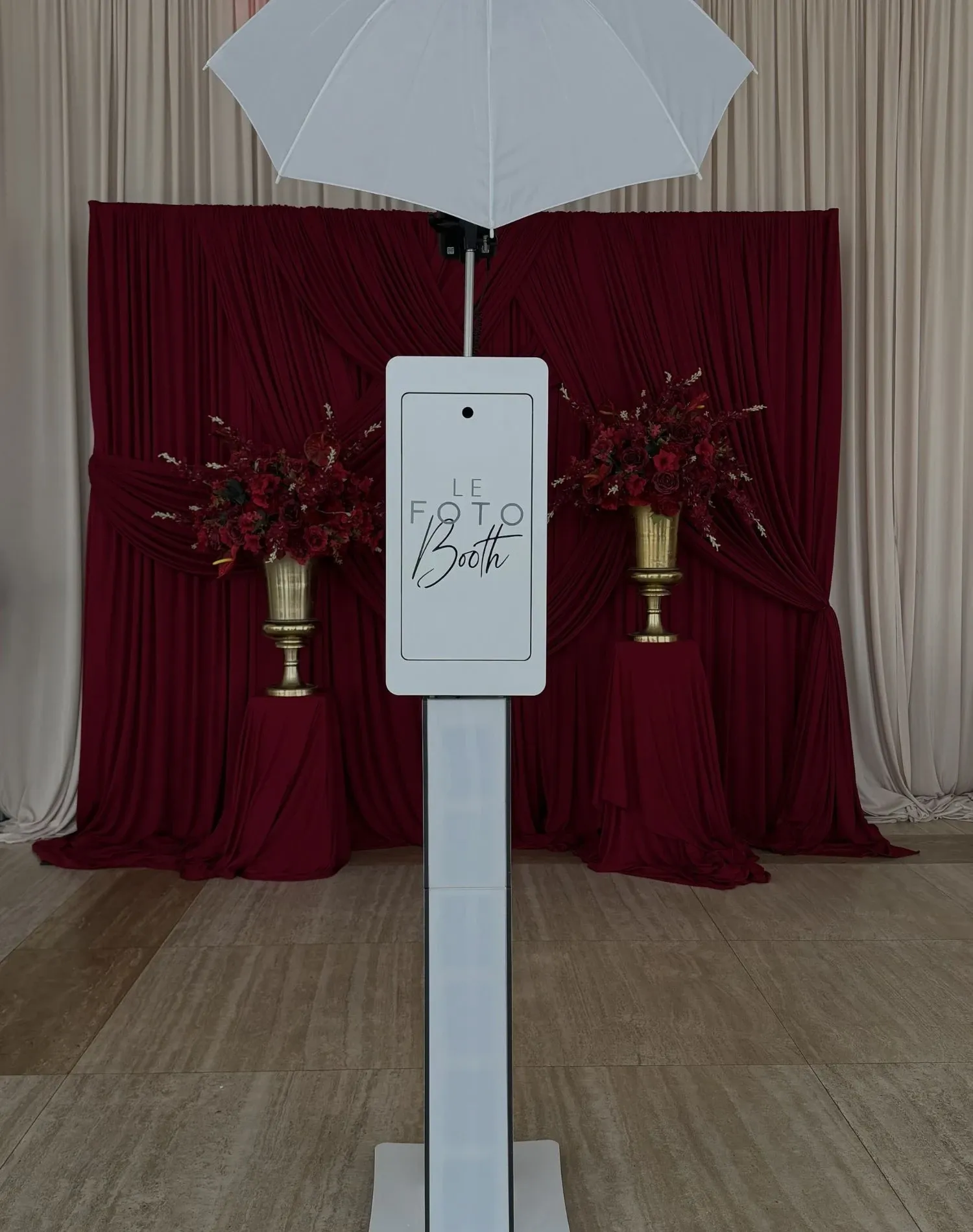 The image shows a sleek, modern photo booth setup by Le Fotobooth, categorized under photo booth vendors for weddings, featured on the Mariage Elegant, Canada website. The photo booth is elegantly designed with a simple white structure and a digital screen displaying the vendor's name. It is positioned against a backdrop of rich burgundy drapes, adding a touch of sophistication. Two large, ornate golden vases filled with deep red flowers flank the booth, enhancing the romantic and luxurious wedding atmosphere.