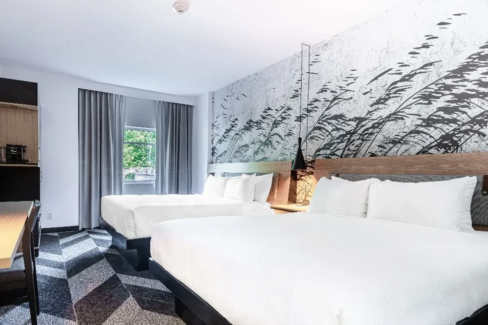 The image shows an elegantly designed guest room at the Doubletree par Hilton Québec Resort, a premier wedding venue. The room features two large, neatly made beds with crisp white linens, perfect for accommodating wedding guests. A sophisticated accent wall with a nature-inspired design adds a touch of modern elegance. Natural light pours in through a window with gray curtains, creating a warm and inviting atmosphere. This setting highlights the resort's commitment to providing comfortable and stylish accommodations for weddings and special events.