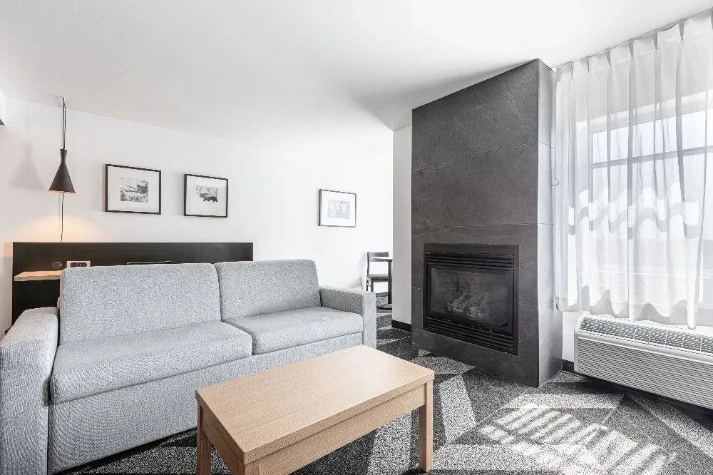 The image showcases a modern and elegantly designed room at Doubletree par Hilton Québec Resort, a premier choice in the wedding venues category. The room features a comfortable grey sofa in front of a sleek, contemporary fireplace, providing a warm and inviting atmosphere. Natural light streams through sheer white curtains, subtly illuminating the space. Clean lines and minimalist decor, including framed artwork and a simple wooden coffee table, enhance the sophisticated ambiance. This venue offers a stylish and cozy setting ideal for wedding preparations or hosting intimate gatherings.