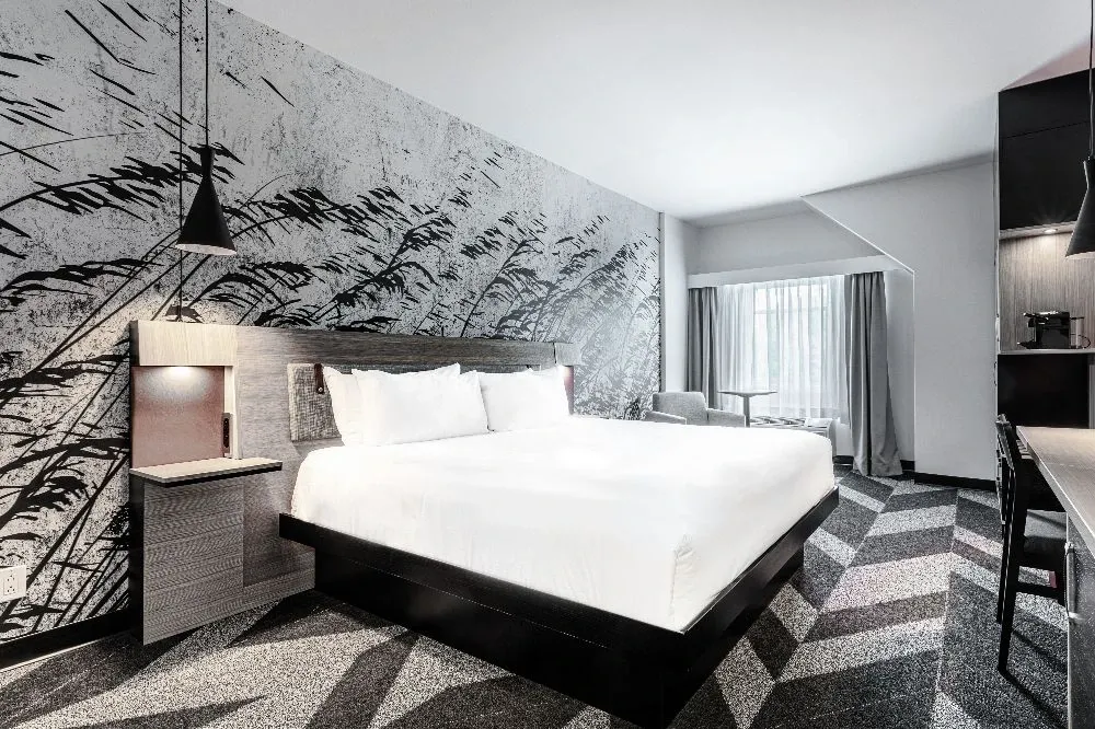 The image shows a modern and stylish guest room at the Doubletree par Hilton Québec Resort, a wedding venue listed under the category of Wedding Venues on Mariage Elegant, Canada. The room features a large, comfortable king-sized bed with white linens, set against a striking abstract mural wall. On each side of the bed, sleek bedside tables with hanging pendant lights add a touch of elegance. The floor is carpeted in a geometric pattern, contributing to the contemporary aesthetic. In the background, a large window lets in natural light, partially covered by light-colored curtains, creating a welcoming and cozy ambiance perfect for accommodating wedding guests.