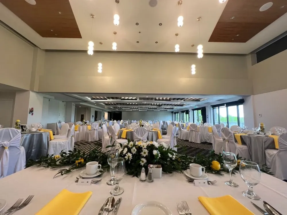 The image displays a beautifully set wedding venue at the Doubletree par Hilton Québec Resort, perfectly fitting the Wedding Venues category. The spacious room is elegantly decorated with round tables draped in white tablecloths, each adorned with a centerpiece made of lush greenery and white flowers accented with yellow blooms. Chairs are covered with white fabric and tied with bows, complementing the table settings. The ceiling features modern light fixtures, and large windows line one side of the room, allowing natural light to illuminate the space. The arrangement exudes a sophisticated and inviting atmosphere, ideal for a wedding celebration.