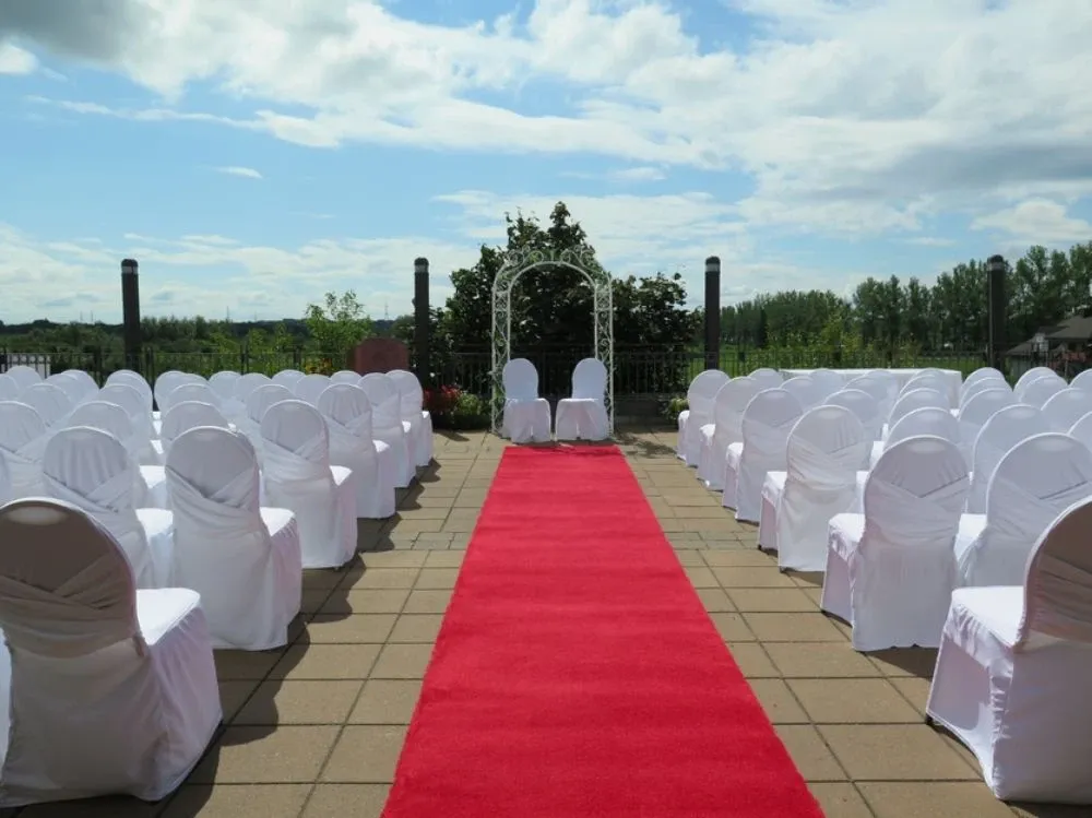 The image shows a beautifully arranged outdoor wedding venue at Doubletree par Hilton Québec Resort, part of the wedding venues category. A red carpet aisle is flanked by rows of white, elegantly covered chairs leading to a decorative white arch adorned with greenery. The setting overlooks a lush landscape under a partly cloudy sky, creating a serene and picturesque environment for a wedding ceremony.