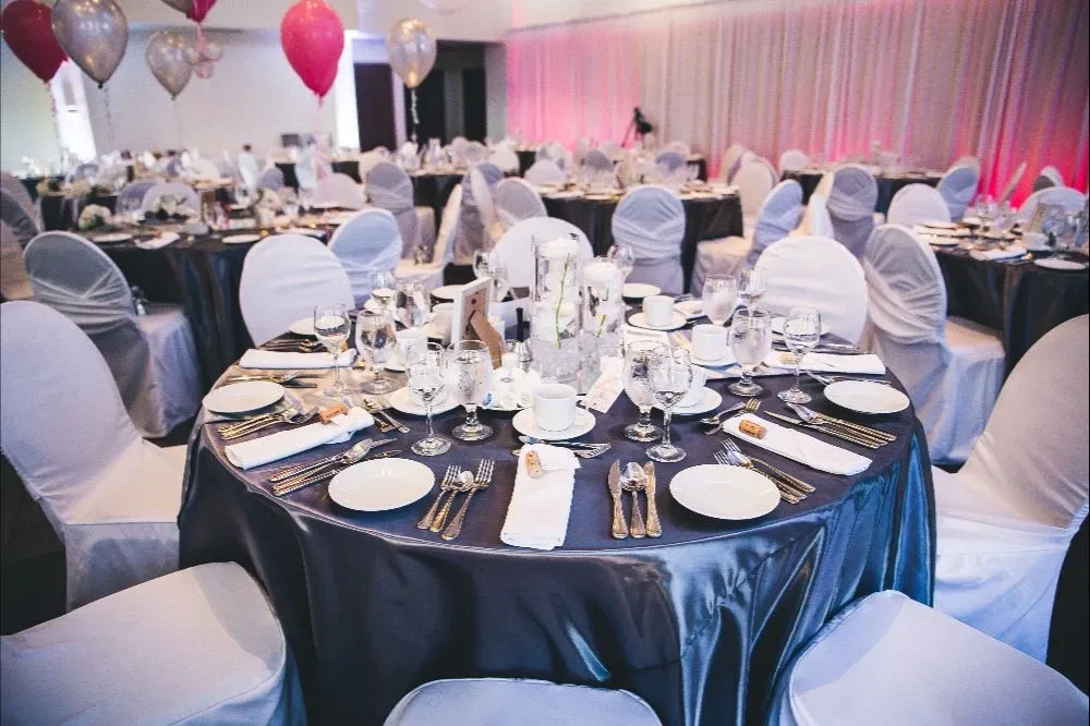 The image showcases a beautifully arranged wedding venue at the Doubletree par Hilton Québec Resort, categorized under wedding venues on the Mariage Elegant site. The setting features elegantly draped round tables with white cloths and dark satin overlays, each adorned with neatly arranged place settings. Fine cutlery, glassware, and simple yet sophisticated floral centerpieces are meticulously placed to create an inviting and upscale dining atmosphere. In the background, white and red balloons add a festive touch, while subtle pink lighting offers a warm ambiance. The room is spacious, providing an elegant setting ideal for weddings.