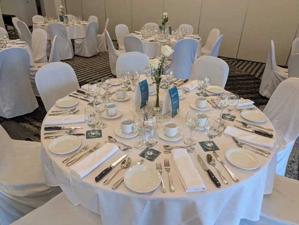 The image shows an elegantly set dining table prepared for a wedding reception at the Doubletree par Hilton Québec Resort, categorized under Wedding Venues. The round table is covered with a crisp white tablecloth and is surrounded by chairs draped in matching white covers. The place settings include pristine white china, polished silverware, neatly folded white napkins, and glassware, reflecting the attention to detail expected at upscale wedding venues. In the center of the table, a simple floral arrangement of white flowers adds a touch of elegance, accompanied by small cardholders displaying menu or guest information, enhancing the sophisticated ambiance. The surrounding tables are identically arranged, indicating a well-coordinated event setup suitable for weddings.