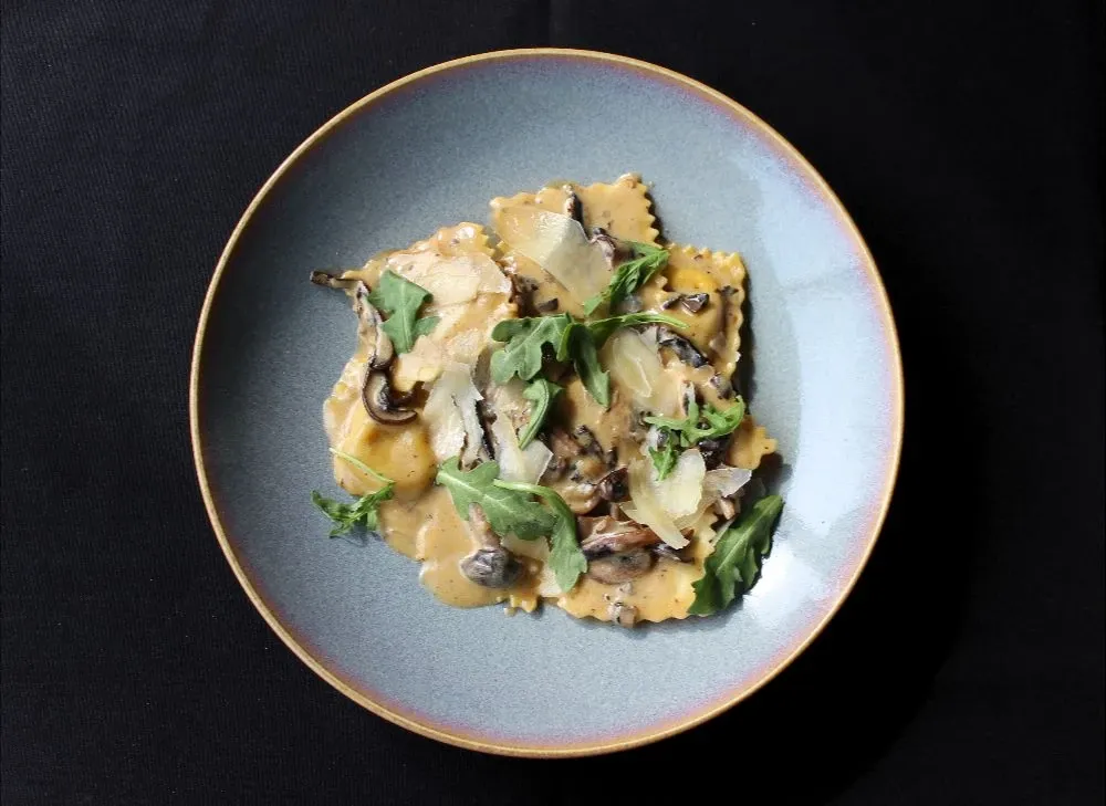 A dish of gourmet ravioli is elegantly presented on a round, light blue plate against a dark background. The ravioli is topped with fresh arugula, thinly sliced mushrooms, and shavings of parmesan cheese. The dish is artfully arranged, showcasing a blend of textures and colors that are visually appealing. This culinary offering is from the Doubletree par Hilton Québec Resort, a venue known for exquisite dining experiences making it an ideal choice for wedding receptions in the Wedding Venues category.