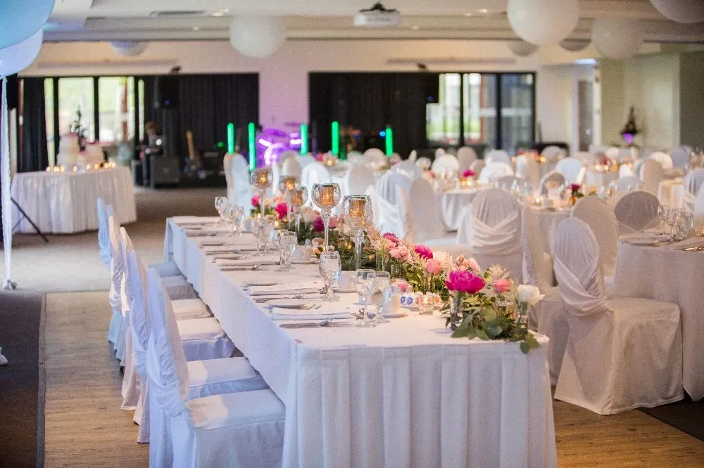 The image showcases a beautifully decorated wedding venue at Doubletree par Hilton Québec Resort, part of the wedding venues category. The spacious hall is elegantly arranged with long tables draped in crisp white linens and lined with matching white covered chairs. Delicate pink and white floral arrangements adorn the tables, accompanied by glass votive candles and sparkling glassware, creating a romantic atmosphere. Ambient lighting and large windows allow natural light to accentuate the sophisticated setting, ideal for a memorable wedding reception.