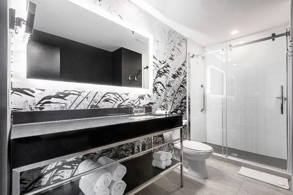 The image showcases an elegant bathroom at the Doubletree par Hilton Québec Resort, under the wedding venues category for Mariage Elegant, Canada. The bathroom features a modern design with a large, backlit mirror above a sleek black countertop. Below the counter, neatly rolled plush white towels are visible. The shower area is enclosed with a clear glass door, and the walls display artistic black and white leaf patterns, adding a sophisticated touch to the space. This luxurious bathroom setting is ideal for wedding preparations or guest accommodations.
