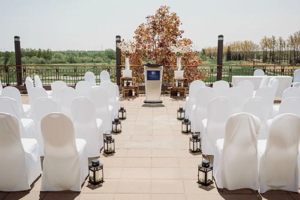 The image depicts an elegantly arranged outdoor wedding venue at the Doubletree par Hilton Québec Resort, part of the wedding venues category on Mariage Elegant, Canada. Rows of white chair covers are neatly aligned, facing a podium adorned with the hotel's emblem, set up for a ceremony. The aisle is flanked by stylish black lanterns, adding a touch of sophistication to the setting. In the background, a vibrant autumn tree provides a picturesque natural backdrop, with views of green landscapes stretching into the distance, creating a serene and inviting atmosphere for a memorable wedding event.