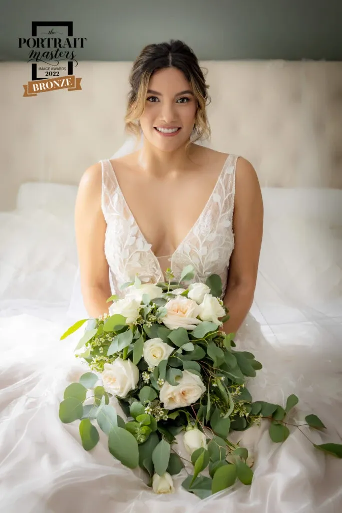 A bride in a detailed, elegant wedding gown is sitting on a bed. She is holding a bouquet of white and peach roses, accented with eucalyptus leaves. Her hair is styled in loose waves. The image includes a badge for 'The Portrait Masters Award, Bronze, 2022.' This wedding photography is showcased by PhotoMarc under the Wedding Photography category on Mariage Elegant, Canada.