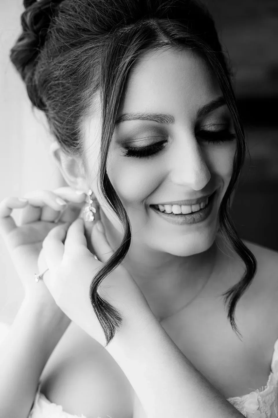 A bride is beautifully captured in black and white as she smiles while adjusting her earrings, showcasing her elegant makeup and intricate hairstyle. This image, provided by the wedding photography vendor PhotoMarc under the wedding category on Mariage Elegant, Canada, highlights the joyful and intimate moments of wedding preparations. Her expression reflects happiness and excitement, adding a personal touch to the special day.