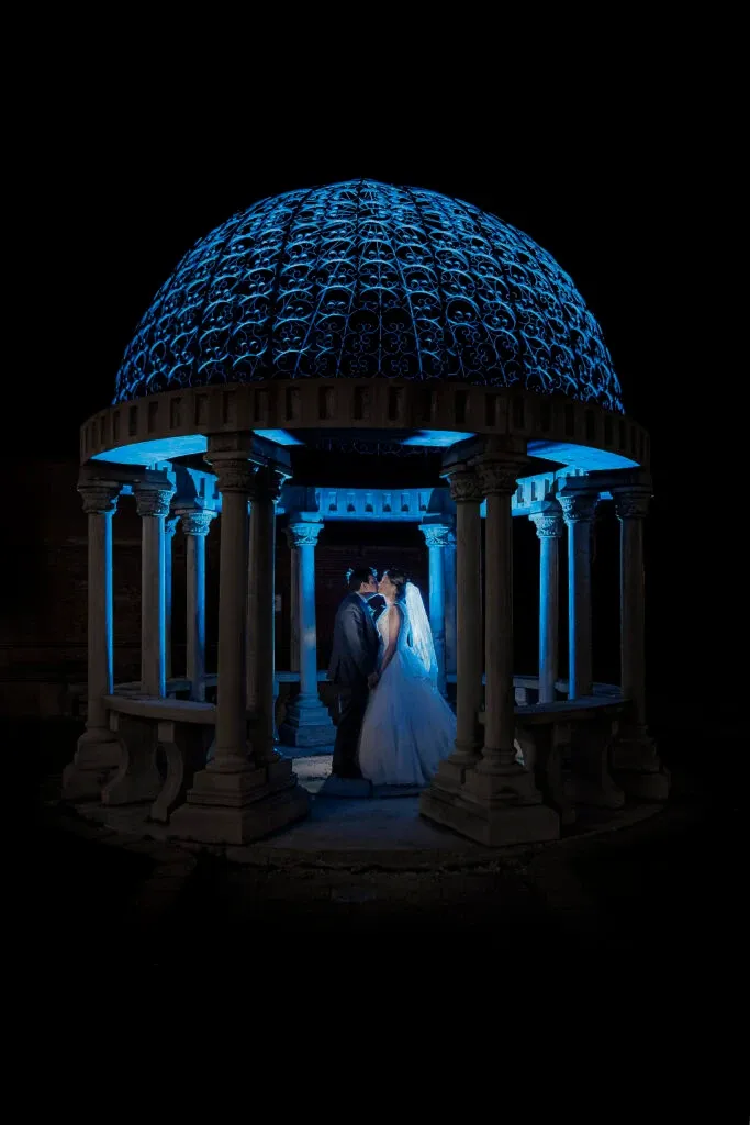 A bride and groom stand intimately beneath a beautifully illuminated, ornate gazebo at night. The structure features intricate architectural details, with a dome cap intricately designed and bathed in a soft blue light, creating a romantic ambiance. The bride is wearing an elegant white gown with a long veil, and the groom is in a classic dark suit. This enchanting scene exemplifies PhotoMarc's expertise in the wedding photography category, capturing timeless moments for weddings in Canada.