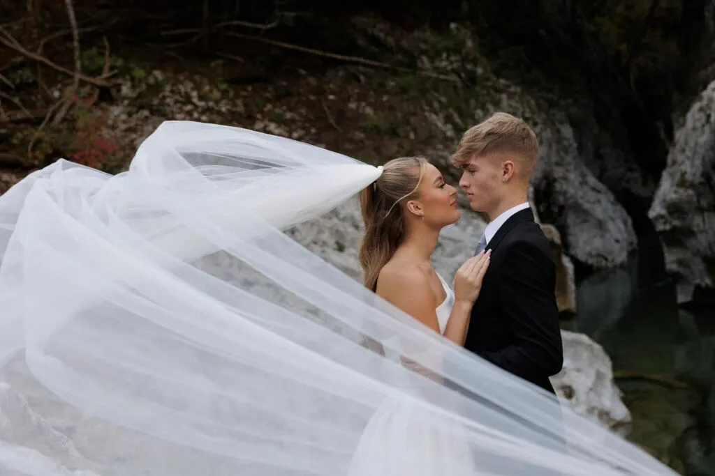 In this image by PhotoMarc, a vendor in the Wedding Photography category for Mariage Elegant, Canada, a bride and groom stand close together in a tranquil setting. The bride's long, flowing veil captures the wind, creating a dramatic effect as it trails behind her. The couple is elegantly dressed, with the bride in a classic white gown and the groom in a sharp black suit. They are positioned near a natural backdrop of rocks and water, adding an element of serene beauty to the scene. The image highlights a tender moment between the couple, perfect for capturing the essence of wedding romance.