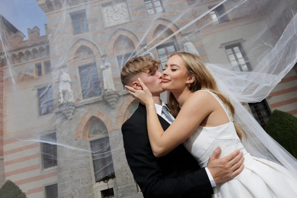 A joyful wedding couple is captured in an affectionate embrace in front of an elegant, historic stone building adorned with statues and arched windows. The bride, in a flowing white gown, playfully kisses the cheek of her groom, who is dressed in a classic black suit. The bride’s long veil billows gracefully in the breeze, adding a sense of romance and movement to the scene. This moment, captured by PhotoMarc, exemplifies their expertise in wedding photography, showcasing their ability to capture intimate and meaningful moments. This image is featured in the wedding photography category for Mariage Elegant in Canada, illustrating the vendor’s skill in creating timeless wedding memories.