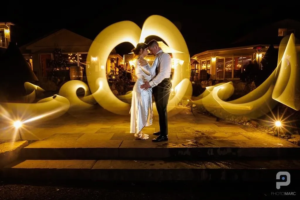 A newlywed couple stands closely together in a romantic embrace, illuminated by warm, ambient lighting in an outdoor setting. The bride wears an elegant, shimmering white gown, while the groom is dressed in a classic black tuxedo. Behind them, a stunning display of light art swirls gracefully, creating a captivating backdrop perfect for a memorable wedding moment. Soft, glowing lights enhance the intimate atmosphere, reflecting the couple's joyous celebration. This image by PhotoMarc, a vendor in the Wedding Photography category at Mariage Elegant, Canada, showcases the artistry and emotional depth captured at weddings.