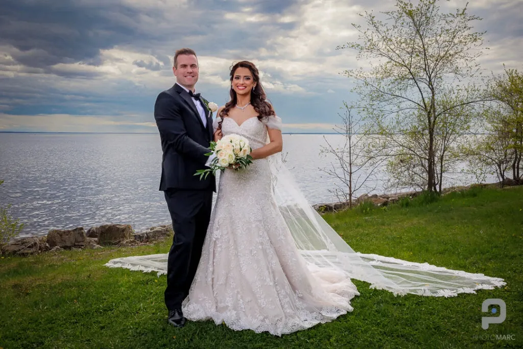 A newly married couple stands on a lush green lawn by a serene lakeside, with a dramatic cloudy sky overhead. The bride is wearing an elegant lace wedding gown and holding a bouquet of white roses, while the groom is in a classic black tuxedo. The peaceful water and natural scenery create a romantic backdrop, perfect for capturing the special day. Photo taken by PhotoMarc, featured in the Wedding Photography category on Mariage Elegant, Canada.