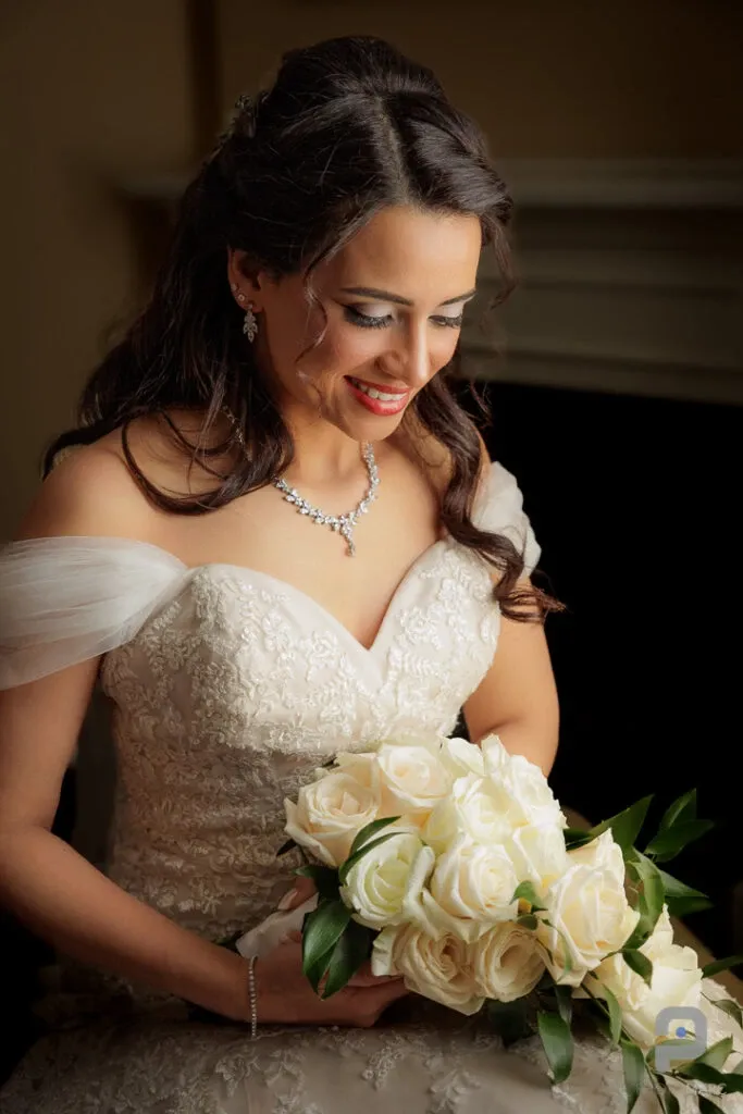 A bride is elegantly posed, holding a bouquet of white roses in this captivating wedding photography shot by PhotoMarc. She is wearing a white, off-the-shoulder lace gown adorned with intricate detailing. Her long, dark hair is styled in soft curls, and she is adorned with delicate jewelry, including a necklace and earrings. The soft lighting enhances her serene expression, creating a romantic and timeless feel, perfectly captured by the vendor in the wedding photography category for Mariage Elegant, Canada.