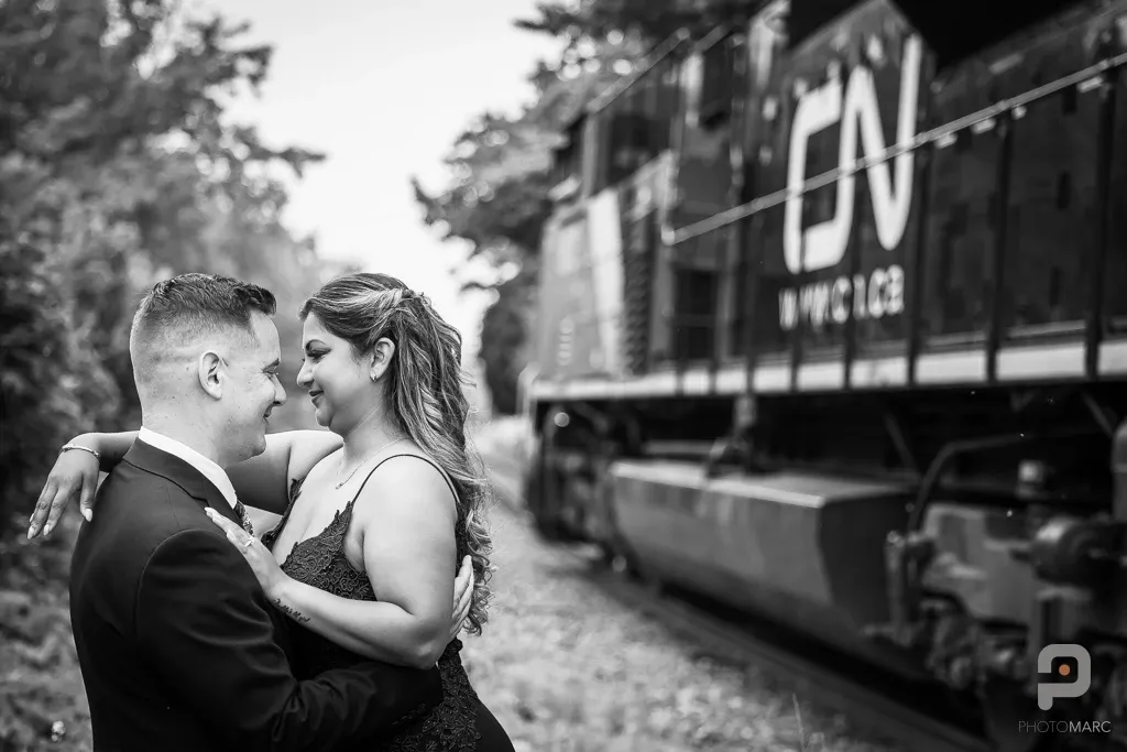 A couple embraces lovingly on their wedding day beside a railroad track in this captivating black and white photograph by PhotoMarc, a vendor specializing in wedding photography. The bride, in a sleeveless lace dress, gazes into the eyes of her groom, who is wearing a stylish suit. The composition beautifully contrasts the romantic moment with the imposing presence of a train passing by, adding a unique and adventurous touch to the wedding setting.
