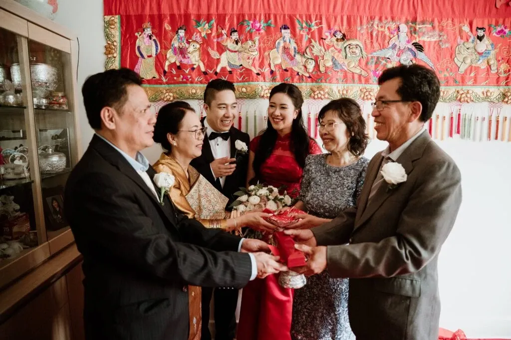 In this vibrant and heartwarming image showcasing KNL Productions in the Wedding Entertainment category for Mariage Elegant, Canada, a group of six elegantly dressed people are gathered in a celebratory moment. Two couples, positioned at the forefront, are engaged in a ceremonial exchange, holding red envelopes—a traditional marriage custom symbolizing luck and prosperity. Behind them, a young couple smiles joyfully; the woman is wearing a striking red dress and holding a beautiful bouquet of white flowers. The setting features a decorative red backdrop adorned with intricate artwork, enhancing the festive atmosphere. The scene captures a mix of cultural tradition and joyful celebration, reflecting the unique services offered by KNL Productions.
