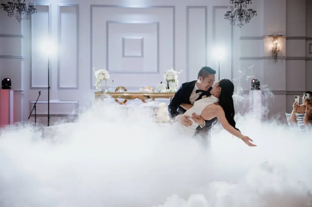 In this elegant wedding photograph from KNL Productions, categorized under Wedding Entertainment on Mariage Elegant, Canada, a newlywed couple is captured in the midst of a graceful dance. The groom dips the bride in a romantic pose, surrounded by ethereal, low-lying fog that adds a dreamlike atmosphere to the scene. The background features a sophisticated, white-paneled wall adorned with chandeliers and floral arrangements on a distant table, enhancing the luxurious setting. Soft, ambient lighting creates a warm and inviting ambiance, perfectly highlighting the couple's joyous moment on their special day.