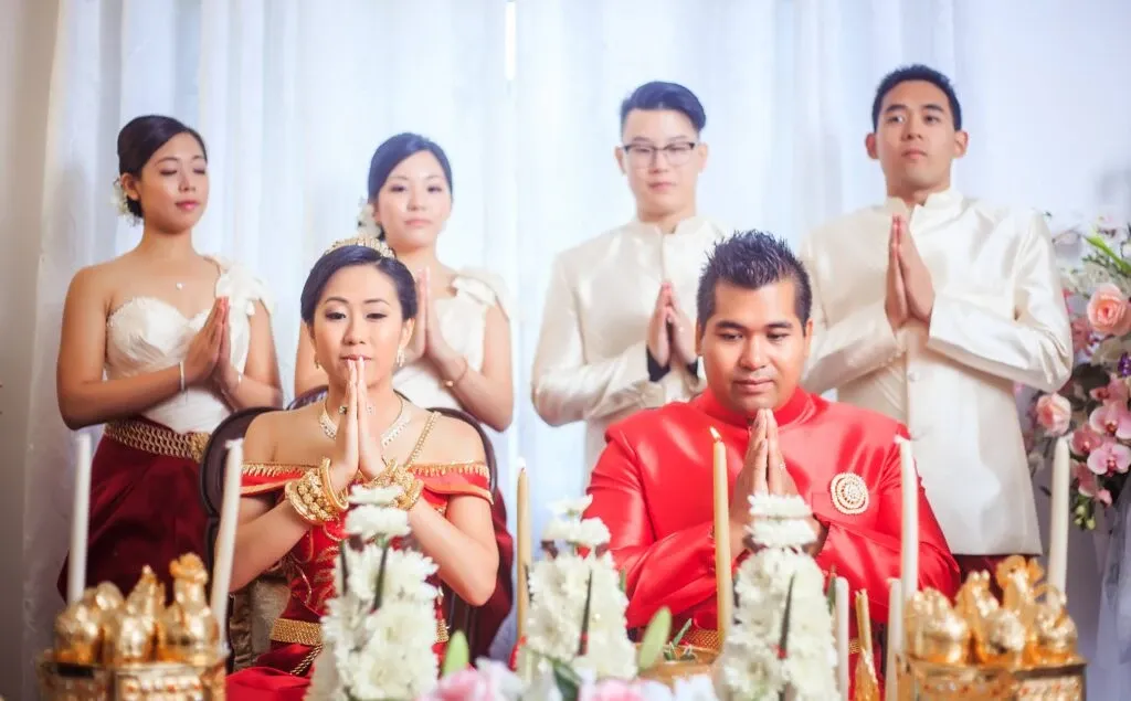 In the image, a bride and groom are seated at the forefront, wearing traditional attire and adorned with gold jewelry, performing a wedding ritual with hands in a prayer position. The bride is clad in a stunning red dress with gold accents, while the groom wears a matching red outfit. Behind them stand four individuals, two men and two women, also dressed in elegant ceremonial clothing, participating in the ritual with joined hands. Floral decorations and ornate golden ornaments add to the celebratory atmosphere. This image represents the wedding entertainment services offered by KNL Productions in the Wedding Entertainment category for Mariage Elegant, Canada.