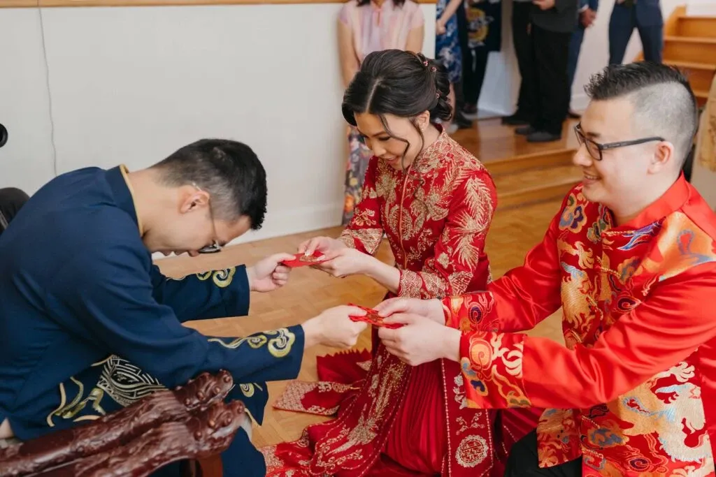 In this image, a wedding couple dressed in traditional Chinese attire participates in a tea ceremony, with the bride and groom kneeling on the wooden floor and serving tea to an elder who is seated. The bride wears a red and gold embroidered dress, while the groom is in a similarly decorated red ensemble. The elder, in a dark blue outfit with subtle designs, bows respectfully. This ceremonial moment is set in a warmly lit room with a few guests observing in the background. KNL Productions has captured this intimate and culturally significant scene, highlighting their expertise in wedding entertainment for Mariage Elegant in Canada.