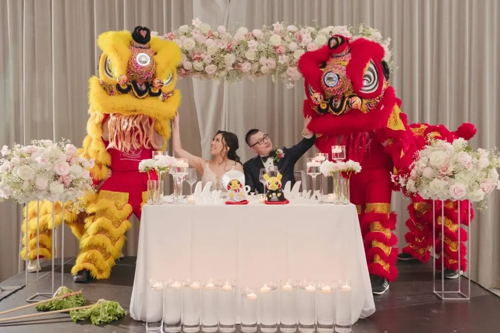 The image features a beautifully decorated wedding reception, highlighting KNL Productions in the Wedding Entertainment category. A couple is seated at a table adorned with elegant floral arrangements and candles in glass holders. The background showcases a stunning floral arch made of pink and white roses. Two performers in vibrant yellow and red lion dance costumes, part of a traditional Chinese lion dance, stand on either side of the couple, adding a cultural touch to the celebration. This setup, presented by KNL Productions, exemplifies a blend of cultural tradition and wedding elegance, making it a memorable part of the wedding festivities.