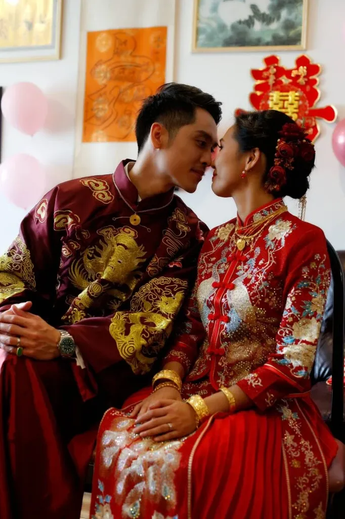 A couple wearing traditional wedding attire sit closely together, facing each other with their foreheads touching in a moment of intimacy. The groom is dressed in an embroidered maroon jacket featuring a golden dragon design, while the bride wears a detailed red dress with intricate gold patterns. Both are adorned with gold jewelry, and the bride has her hair styled with decorative red floral accents. In the background, festive red and gold decorations, including a double happiness symbol, are visible alongside soft pink balloons. This image represents KNL Productions in the Wedding Entertainment category for Mariage Elegant in Canada, highlighting their ability to capture culturally significant moments for weddings.