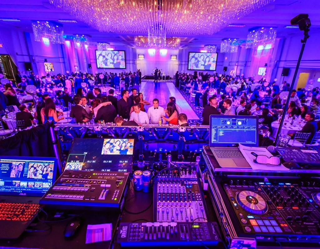 The image showcases a vibrant wedding reception setting provided by KNL Productions, a vendor specializing in wedding entertainment. The scene is illuminated with a captivating purple and blue lighting ambiance, characteristic of a lively celebration. At the forefront, there is a comprehensive DJ and sound mixing setup, featuring mixers, turntables, and laptops with sound equipment ready for the evening's entertainment. Beyond the setup, numerous elegantly dressed guests are seated at round tables adorned with white tablecloths, socializing and enjoying the event. The background displays large screens projecting images of the newlyweds, complementing the grand chandelier-lit ballroom. The image captures the professional and energetic atmosphere KNL Productions brings to wedding celebrations.
