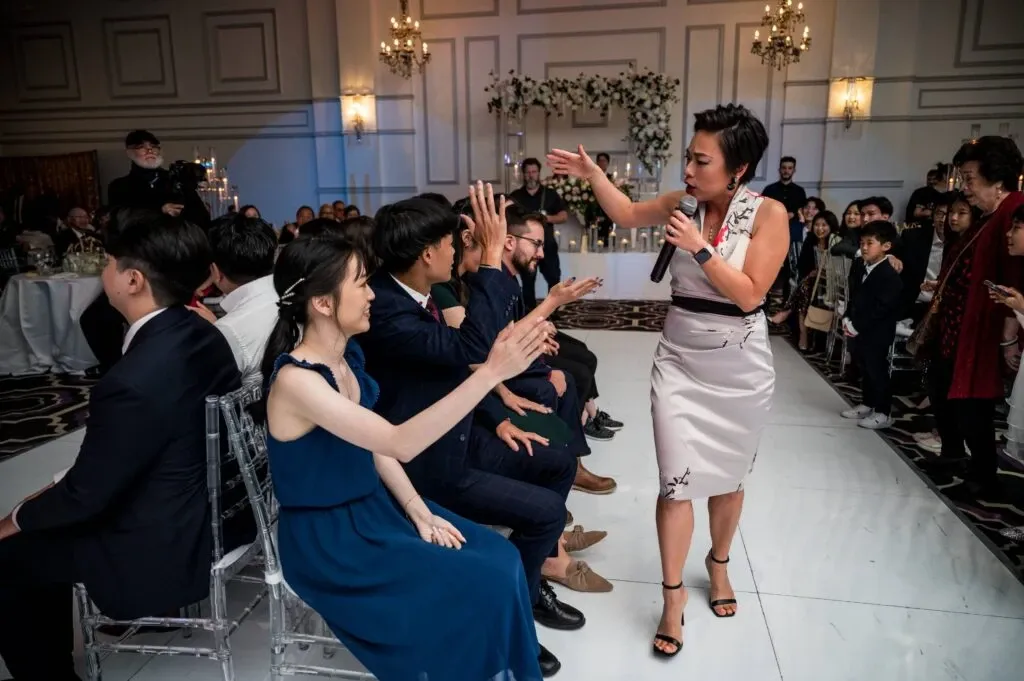 In an elegant wedding reception setting, a professional from KNL Productions, specializing in wedding entertainment, energetically engages with seated guests. The entertainer, dressed in a stylish outfit, interacts with the guests who are smiling and participating eagerly. The beautifully decorated venue features chandeliers and floral arrangements in the background, contributing to the joyous atmosphere. This setup exemplifies the lively and interactive services offered by KNL Productions at wedding events.