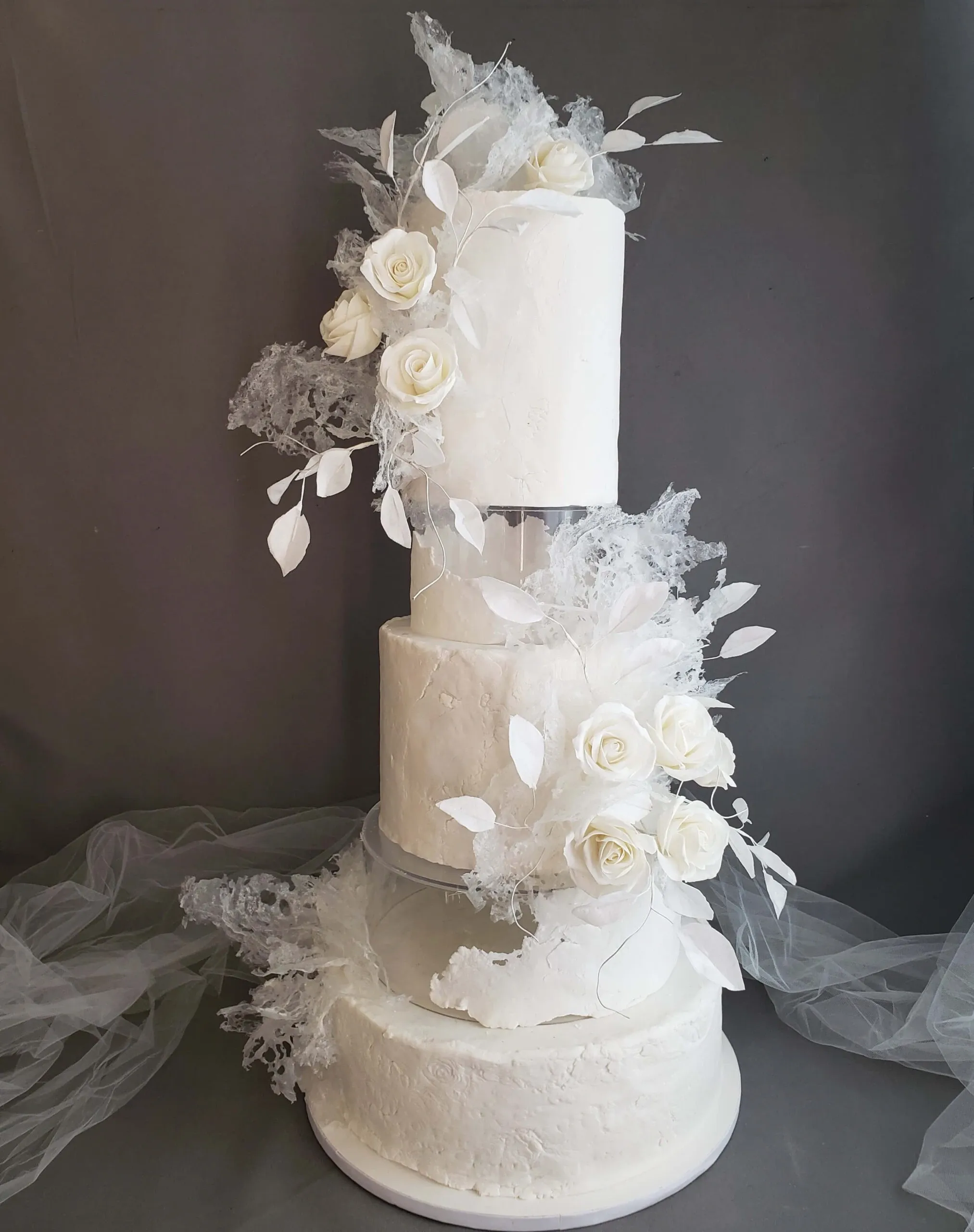 This image features a stunning three-tier wedding cake by Patisserie Artisanale Kalina, showcased in the wedding cakes category. The design boasts a smooth white fondant finish, elegantly adorned with delicate white roses and artistic sugar leaves. Each tier is separated by a clear acrylic layer, adding a modern touch and giving the illusion that the tiers are floating. The cake is further decorated with translucent edible sugar structures, creating an ethereal, sophisticated look perfect for an elegant wedding setting.