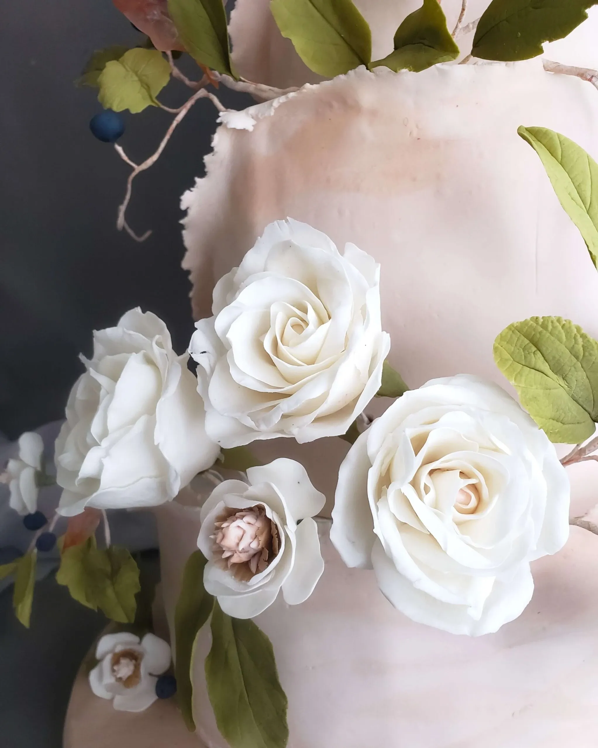 This image showcases a beautifully crafted wedding cake from Patisserie Artisanale Kalina, categorized under Wedding Cakes on Mariage Elegant, Canada. The cake is adorned with intricate, life-like white sugar roses and delicate green leaves, adding a touch of elegance and sophistication. The detailed artistry in the floral arrangement highlights the vendor's skill in creating exquisite wedding confectionery. The background is a soft, neutral tone, emphasizing the cake's decorative elements, ideal for a romantic wedding setting.