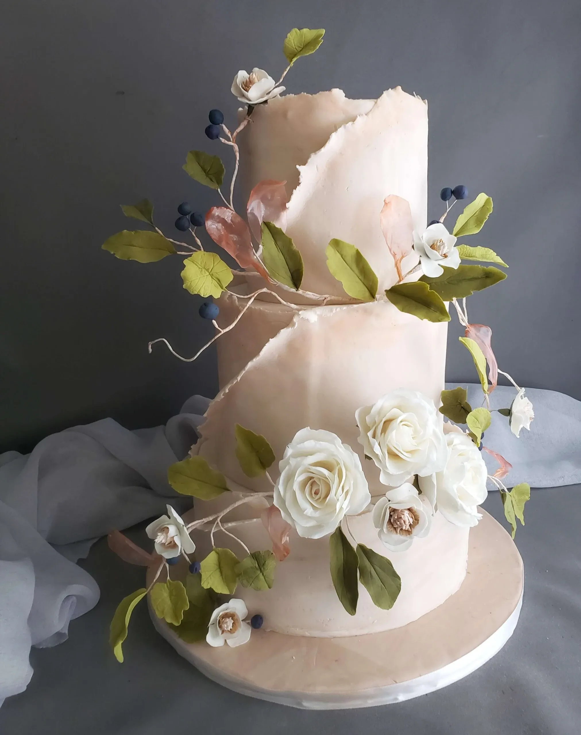 This image showcases an elegant, two-tiered wedding cake crafted by Patisserie Artisanale Kalina, listed in the Wedding Cakes category on Mariage Elegant, Canada. The cake features a sophisticated design with soft blush-colored icing and expertly crafted decorative elements. Delicate white roses made of sugar adorn the base, accented with green leaves and small blue berries, lending a natural and artistic flair. Pink translucent leaves add a touch of whimsy, enhancing the organic aesthetic. The cake sits against a gray fabric backdrop, highlighting its refined details and artistic craftsmanship, ideal for a stylish wedding celebration.
