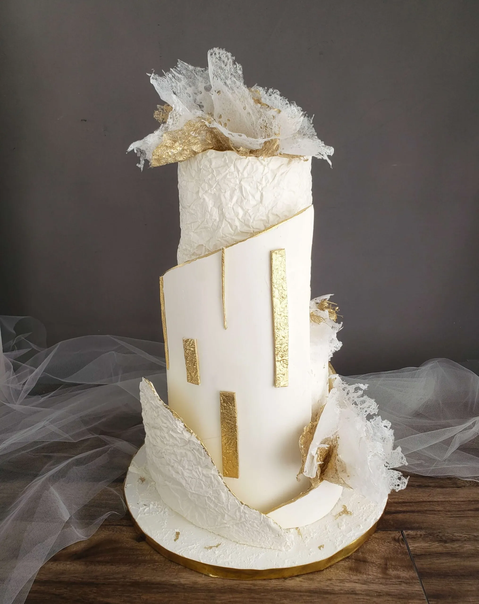 This image showcases a stunning wedding cake by Patisserie Artisanale Kalina, featured in the Wedding Cakes category on Mariage Elegant, Canada. The cake is a tall, cylindrical masterpiece adorned with elegant white fondant that has a textured finish resembling delicate fabric. It is accented with striking vertical gold leaf details, adding a touch of luxury. The cake is further adorned with artistic white wafer paper decorations at the top and base, creating an intricate and ethereal appearance. The cake rests on a simple, gold-edged board, complementing its sophisticated design. The soft gray background enhances the cake's elegant and modern aesthetic, making it an ideal centerpiece for an upscale wedding celebration.