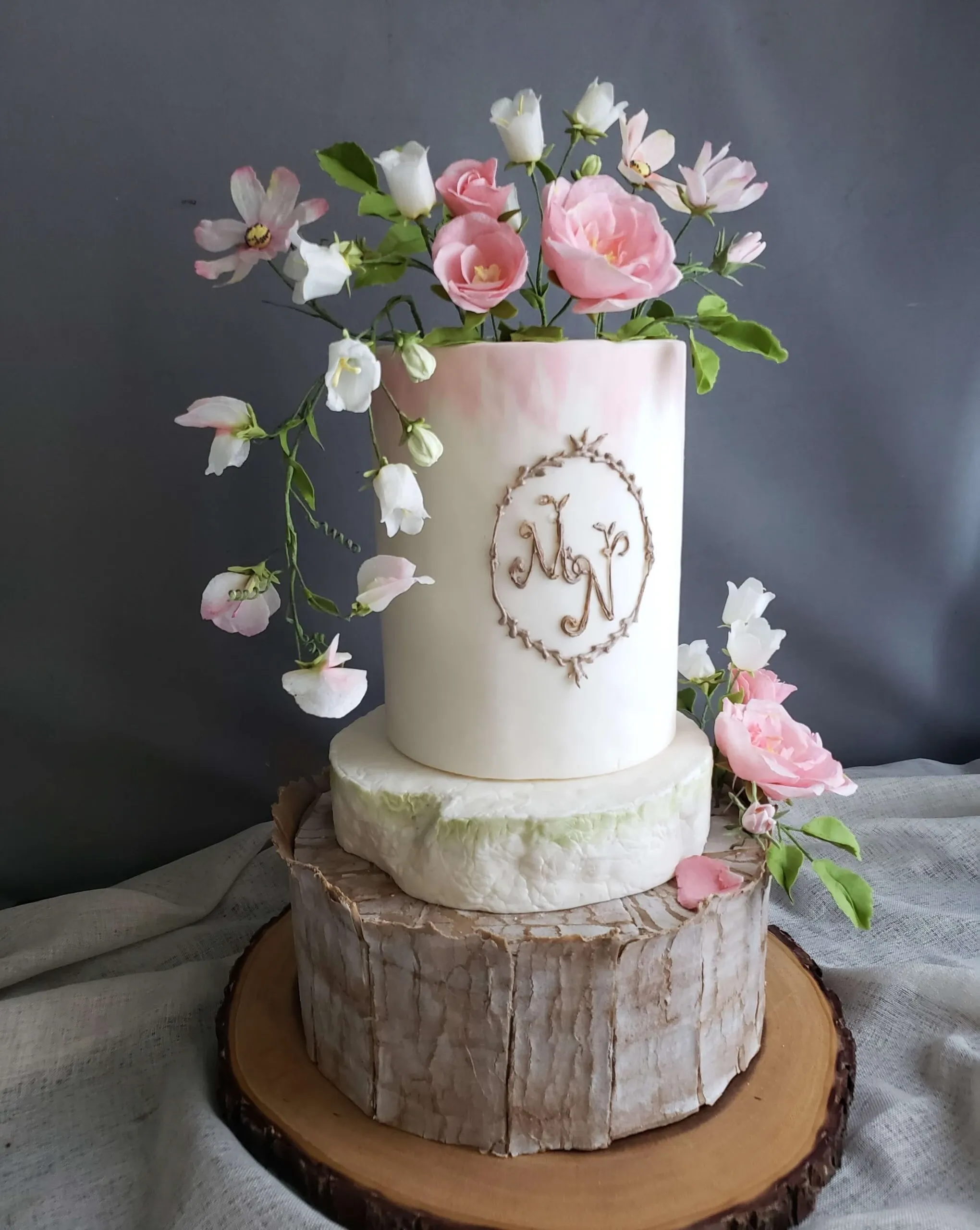 This image showcases an elegant wedding cake crafted by Patisserie Artisanale Kalina, highlighted under the wedding cakes category on the Mariage Elegant site in Canada. The cake features a sophisticated, two-tiered design. The top tier is adorned with delicate pink and white flowers, including roses and small blossoms, creating a natural and romantic atmosphere. The cake is set on a rustic wooden base, adding a charming rustic elegance. A monogram is beautifully inscribed in a golden wreath on the front, contributing to the personalized wedding theme. The overall presentation of the cake reflects the exquisite artistry and dedication to detail typical of Patisserie Artisanale Kalina.