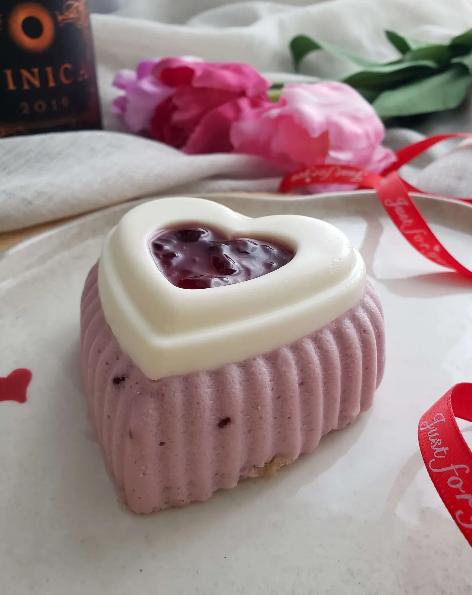 This image shows a beautifully crafted heart-shaped wedding cake by Patisserie Artisanale Kalina, featured in the Wedding Cakes category. The cake has a textured light purple base and is elegantly topped with a white chocolate heart containing a rich red berry compote. It is set against a soft background with pink flowers and decorative red ribbons, enhancing the romantic wedding theme. This elegant piece is perfect for adding a touch of sweetness to a sophisticated wedding celebration by Mariage Elegant Canada.