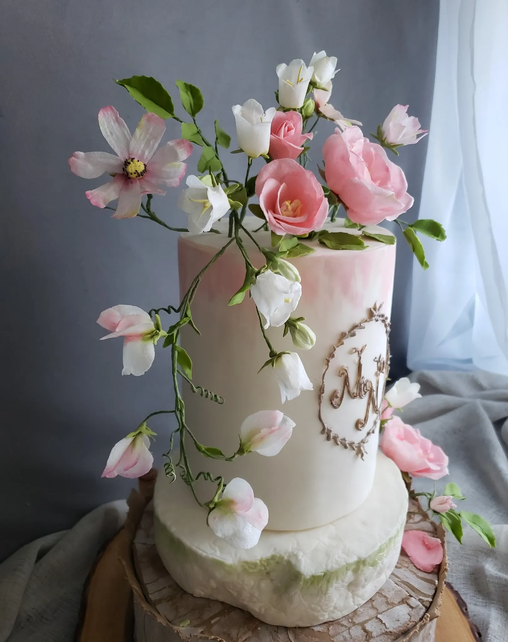 This image showcases a beautifully handcrafted wedding cake by Patisserie Artisanale Kalina, falling under the Wedding Cakes category on Mariage Elegant, Canada. The cake is a two-tiered masterpiece with a smooth, ivory fondant finish. The top tier features an elegant design with the initials 'M' and 'J' intricately piped in gold. Cascading from the top are delicate, lifelike sugar flowers, including pink and white roses and sweet peas, adding a touch of romance and elegance. The cake is displayed on a rustic wooden base, enhancing its charm, with soft natural light seeping through a nearby window.