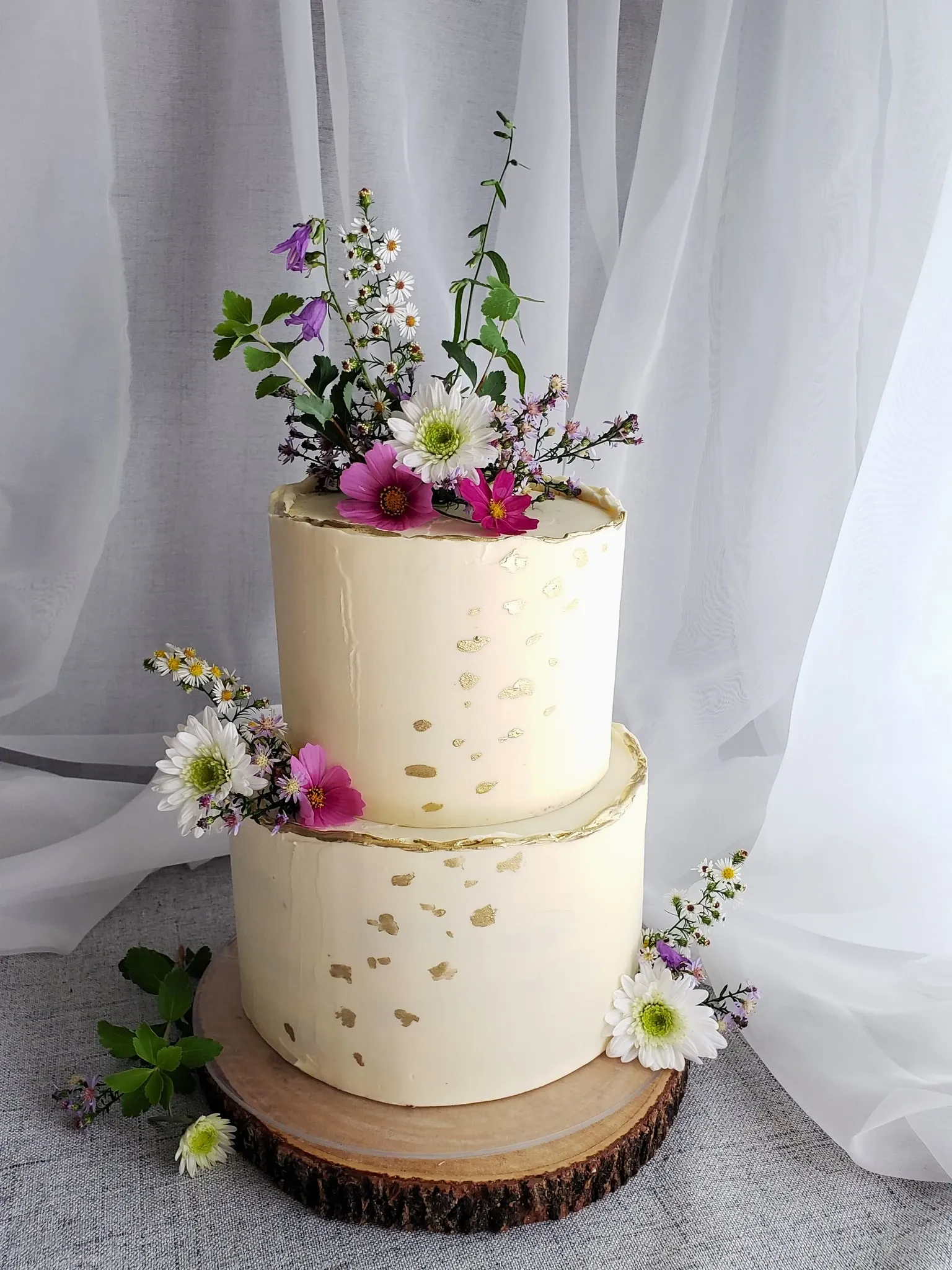 This image features a beautifully crafted wedding cake by Patisserie Artisanale Kalina, under the wedding category of elegant cakes. The two-tiered cake is adorned with a smooth, white buttercream frosting, delicately speckled with gold leaf accents for a touch of luxury. Atop the cake and cascading down its sides are fresh, colorful flowers, including daisies and wildflowers, creating a vibrant and romantic display. The cake is elegantly placed on a rustic wooden stand, enhancing its artisanal and sophisticated appeal, perfect for a refined wedding celebration. The soft, sheer curtain backdrop adds to the overall elegance of the presentation, making it an ideal choice for a stylish wedding reception.