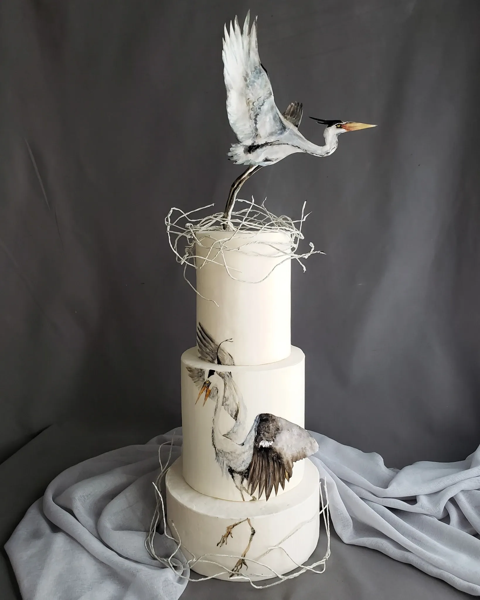 This image features a stunning wedding cake created by Patisserie Artisanale Kalina, under the Wedding Cakes category on Mariage Elegant, Canada. The cake is a three-tiered masterpiece with a smooth, white fondant finish. Delicately sculpted and painted herons adorn the cake, with one heron standing gracefully atop the highest tier, its wings elegantly outstretched, and another depicted mid-flight on the side. Naturalistic elements such as twigs and fine branches complement the intricate design, enhancing the artistic and elegant theme perfect for a sophisticated wedding celebration.