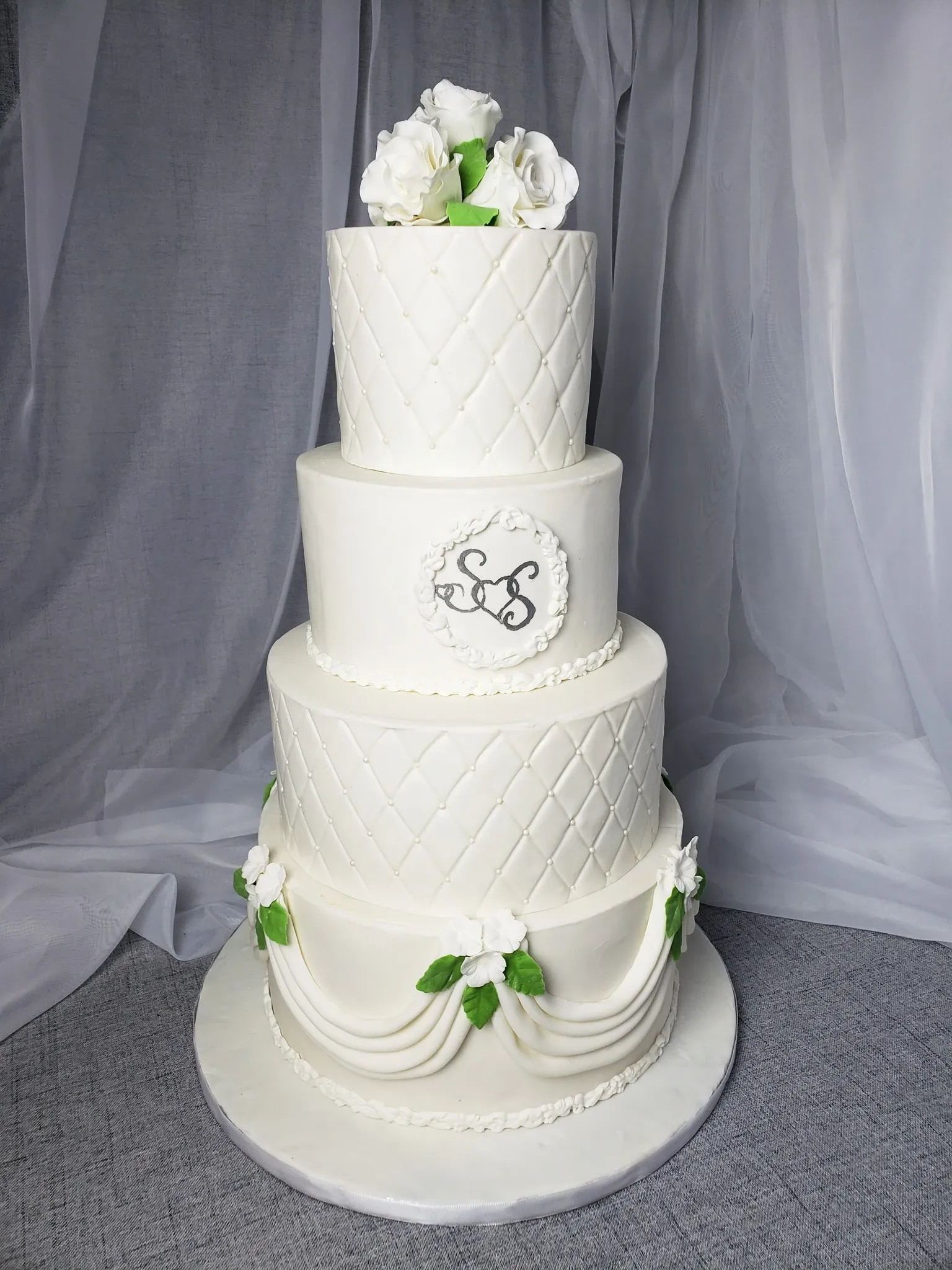 This image features a beautifully crafted wedding cake by Patisserie Artisanale Kalina, prominently displayed under the wedding cakes category. The cake is a stunning three-tier creation, elegantly covered in smooth white fondant with a quilted design on each tier. Delicate white roses with lush green leaves adorn the top and base of the cake, adding a touch of natural beauty. Intricate draping embellishments are also sculpted in fondant, giving the cake a refined and classic appeal. The middle tier features a decorative plaque with interlocking initials, contributing a personalized touch. This sophisticated design embodies the elegance and artistry perfect for a wedding celebration.