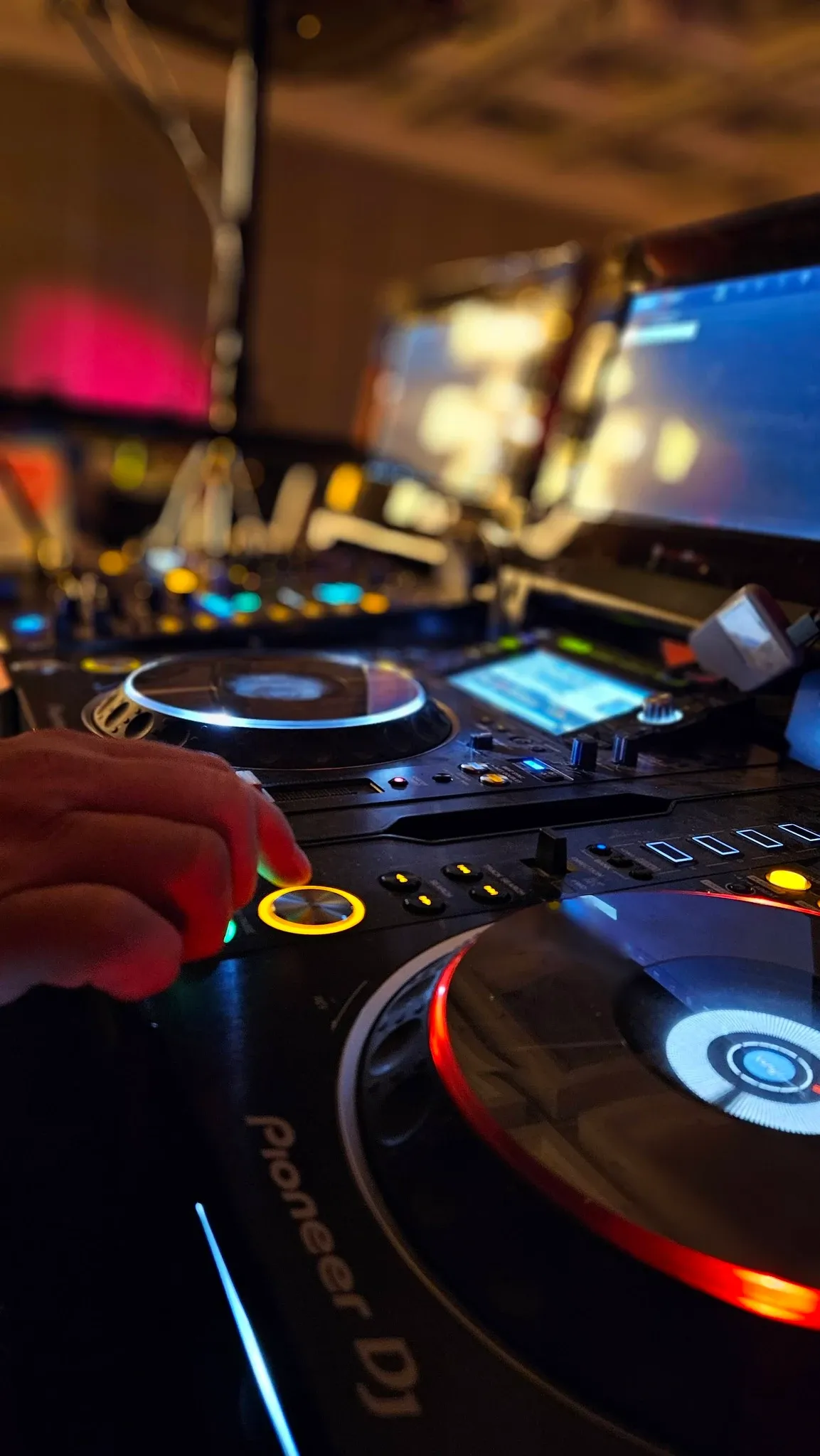 A DJ setup is captured in vibrant detail, featuring a close-up of a hand skillfully adjusting controls on a professional Pioneer DJ mixer, illuminated with colorful LED lights. The background displays softly blurred lights and screens, suggesting a lively and dynamic wedding atmosphere. This image represents Productions Megamix in the Wedding Entertainment category, highlighting their expertise in creating memorable musical experiences for weddings.