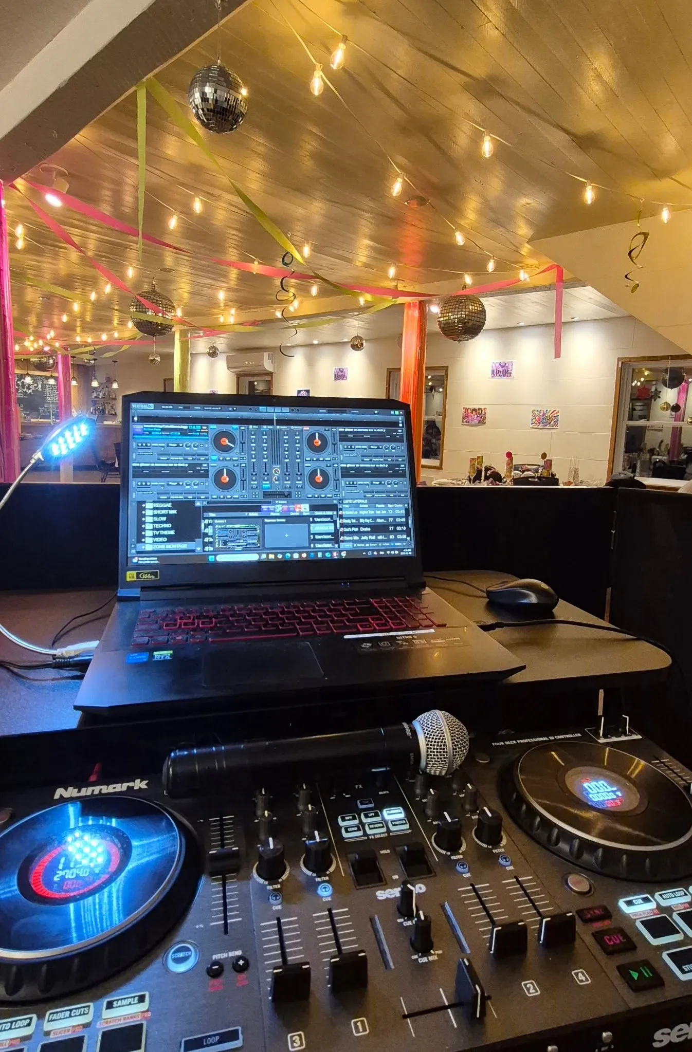 The image shows a vibrant wedding entertainment setup by Productions Megamix, a vendor in the wedding category. A DJ booth is prominently displayed, featuring a Numark DJ controller and a laptop showing digital music mixing software, ready for a dynamic performance. Colorful streamers and sparkling disco balls hang from the ceiling, with warm string lights adding a festive ambiance to the venue. In the background, tables are elegantly set for wedding guests, enhancing the celebratory atmosphere. This setup highlights the lively and professional wedding entertainment services offered by Productions Megamix.