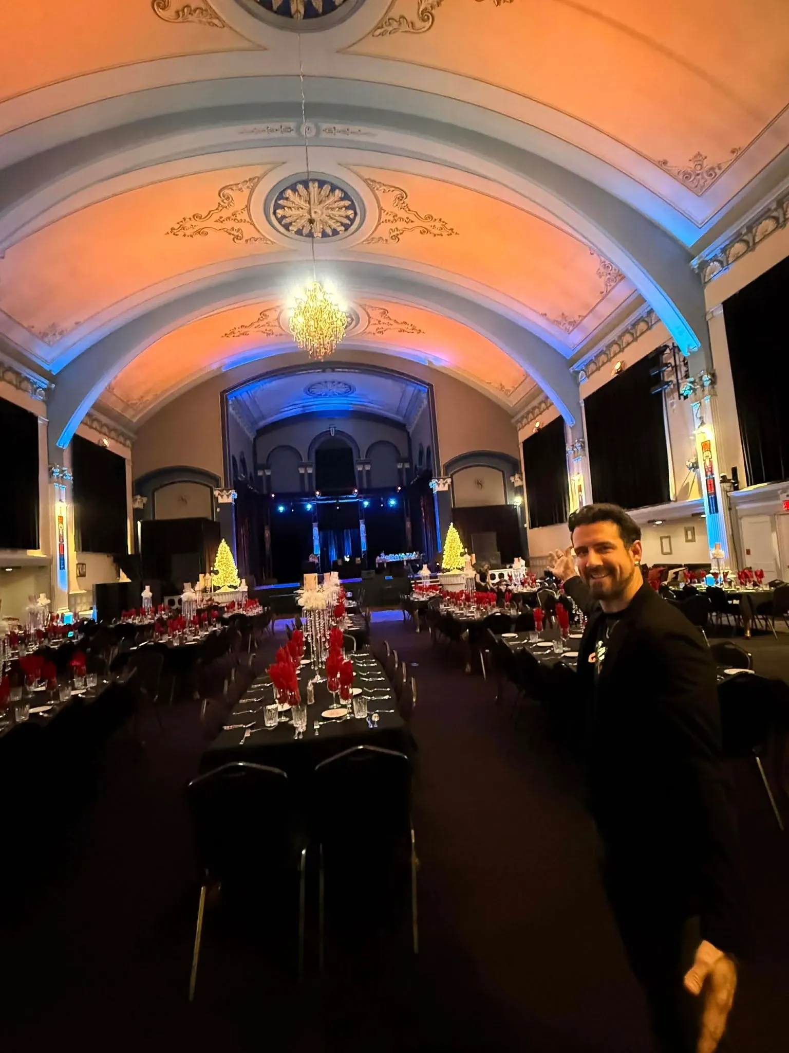 This image showcases a beautifully lit wedding reception hall featuring the work of Productions Megamix, a vendor specializing in Wedding Entertainment. The venue is elegantly decorated with ornate ceiling designs and a stunning central chandelier. Warm lighting enhances the elegant architecture and creates an inviting atmosphere. Rows of neatly arranged tables are adorned with red napkins and black chairs, with festive decorations visible throughout the setting. The foreground includes a person in formal attire, suggesting the lively engagement typical of a wedding celebration.