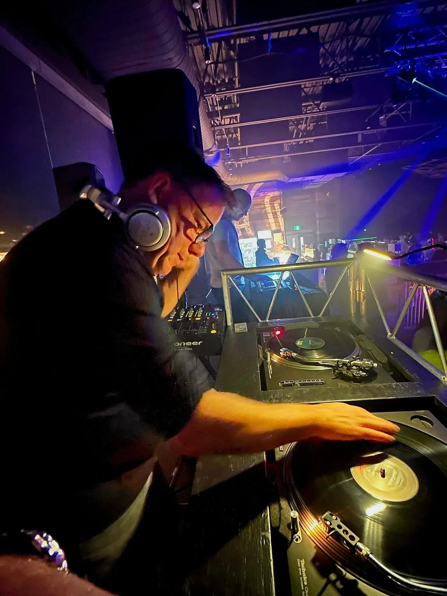 A DJ from Productions Megamix, featured in the wedding entertainment category, is skillfully mixing music on a turntable in a vibrant wedding reception setting. The DJ is wearing headphones and focused on the vinyl records, creating an energetic and engaging atmosphere. The background is filled with colorful lights and a lively crowd, contributing to a festive celebration. This image highlights the dynamic and professional entertainment services provided by Productions Megamix for weddings in Canada.