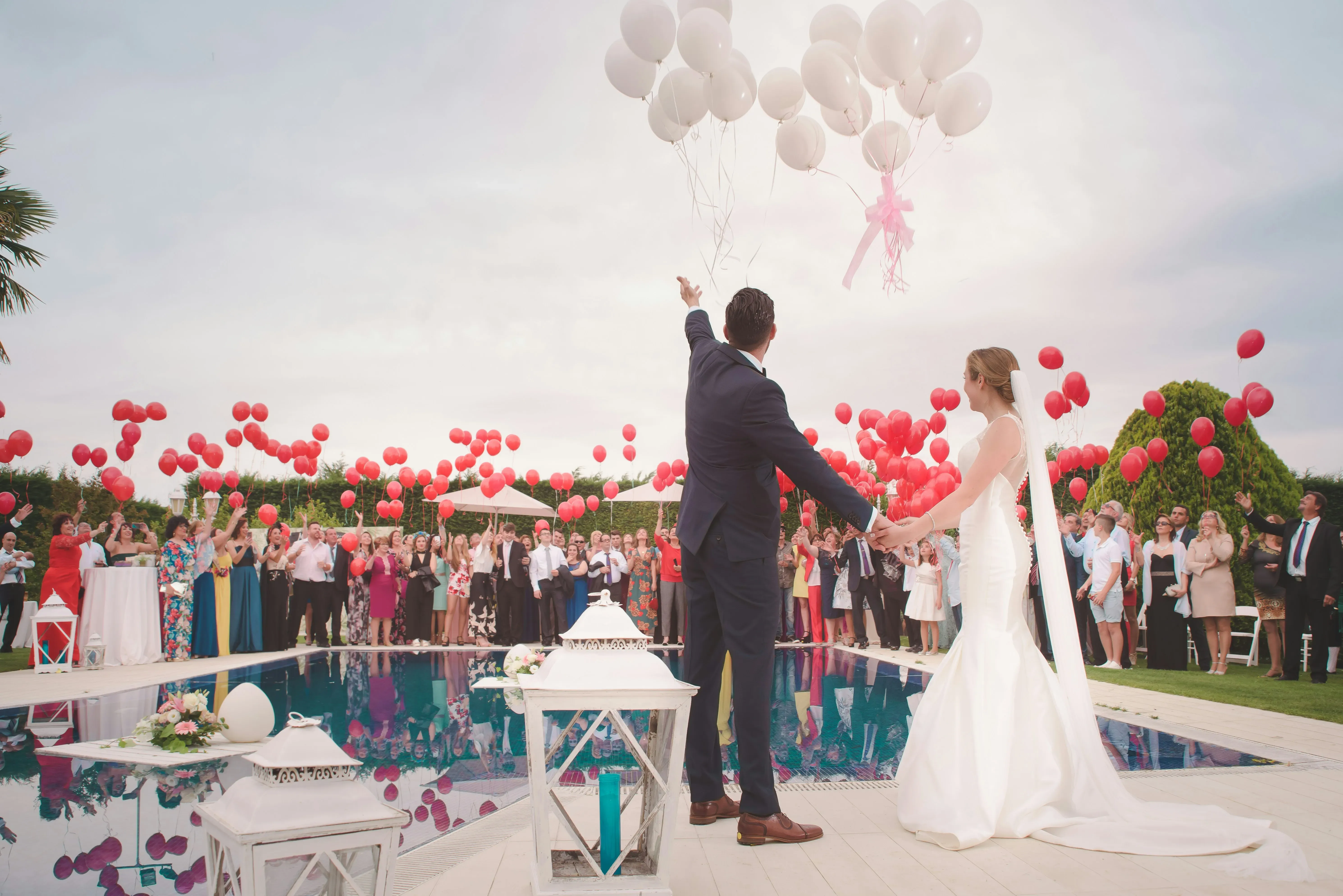 A joyful couple in formal wedding attire stands hand-in-hand beside a shimmering pool, watching white balloons ascending into a partly cloudy sky. The bride in a flowing white gown and the groom in a classic suit engage with their guests, who are gathered around the pool, holding red balloons in celebration. Surrounding the pool area are decorative white lanterns, adding to the elegant ambiance. This image captures a lively and picturesque scene perfect for a destination wedding, showcasing the services offered by Voyage Club Evasion in the category of Destination Wedding on Mariage Elegant, Canada.