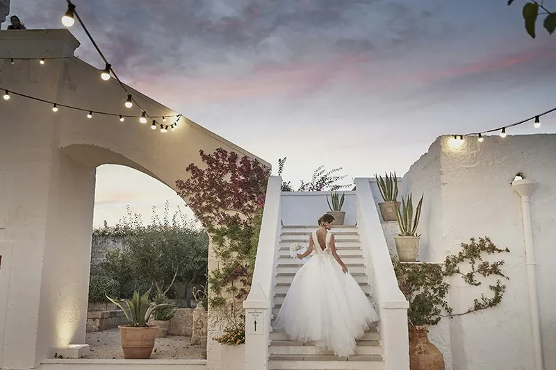A bride in a flowing white gown stands gracefully on a grand staircase outside an elegant white-walled villa, surrounded by lush greenery and potted plants. The evening sky is painted with soft hues of pink and blue, adding a romantic ambiance to the scene. Warm string lights are strung overhead, enhancing the enchanting atmosphere. This image showcases the destination wedding expertise of Voyage Club Evasion, highlighting their ability to create beautiful and memorable settings for special occasions. The exquisite architecture and serene environment make it an ideal location for an unforgettable wedding celebration.