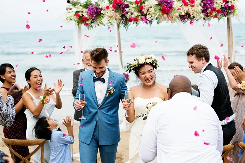 The image shows a joyful couple at their destination wedding, organized by Voyage Club Evasion, under the category of Destination Wedding. The bride, wearing a white gown and a floral crown, and the groom in a light blue suit, are walking down an aisle on a beach, surrounded by cheering guests. Pink petals are being thrown in celebration as they hold glasses, likely filled with champagne. The ocean is visible in the background, alongside a ceremonial arch beautifully decorated with vibrant flowers in shades of pink, purple, and white. This setting exemplifies the elegance of a romantic beach wedding.