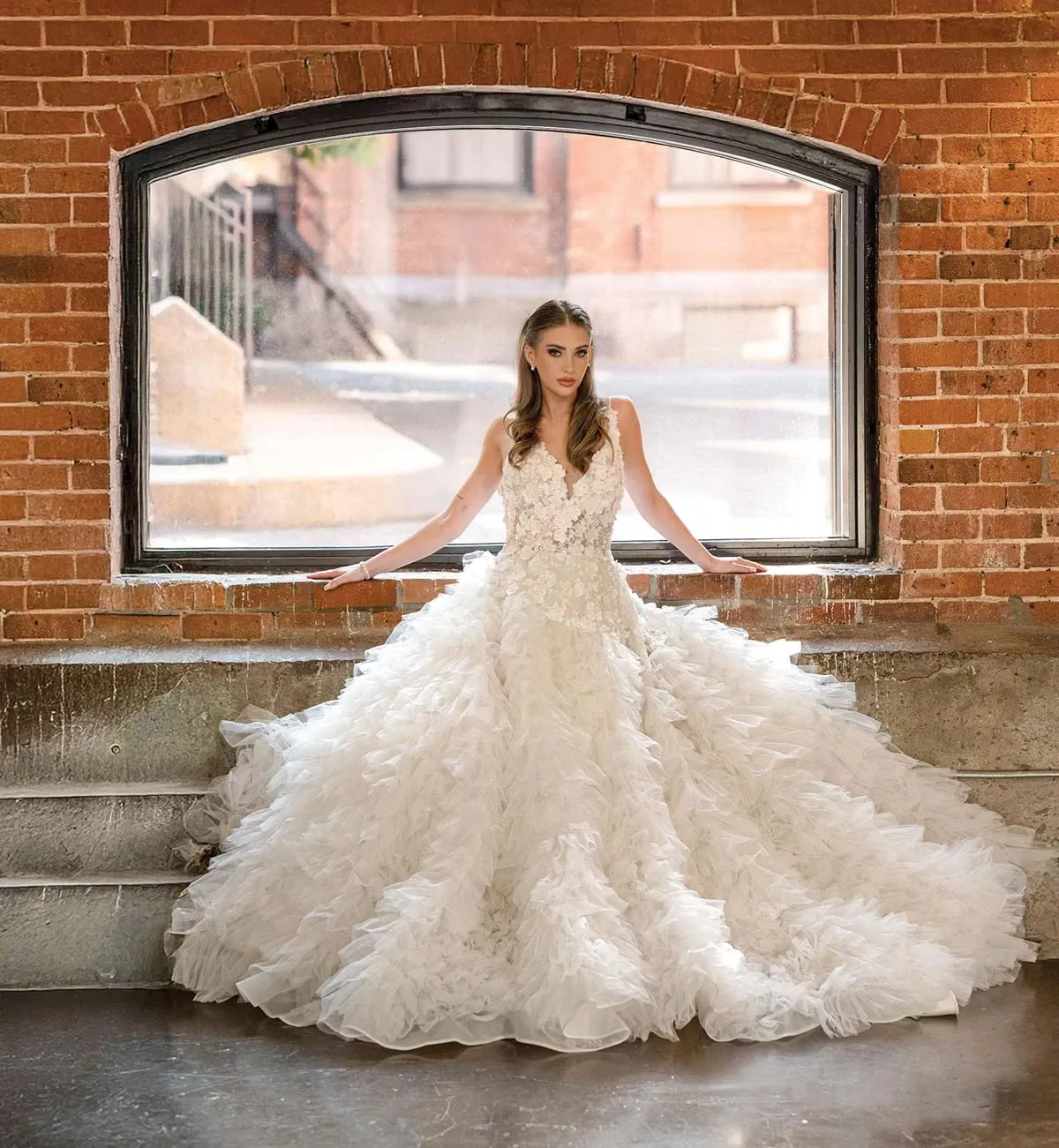 A bride poses elegantly in front of a large, arched window with an exposed brick background. She is wearing a voluminous, ruffled white wedding gown with intricate lace detailing on the bodice, creating a stunning contrast against the rustic setting. Her long hair is styled in soft waves, complementing her natural makeup. The scene is illuminated by natural light streaming through the window, highlighting her graceful posture and the gown's elaborate design. This image is associated with Fusion Beaute, showcasing their expertise in Wedding Beauty for Mariage Elegant in Canada.