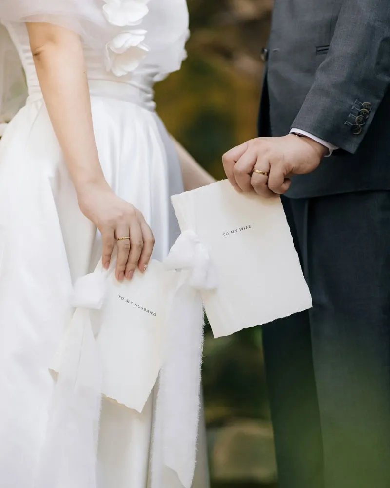 La photo montre un couple de mariés, tenant chacun une carte de vœux élégante. La mariée, vêtue d'une robe blanche ornée de fleurs en tissu, tient une carte avec l'inscription 'To My Husband'. Le marié, portant un costume gris foncé, tient une carte avec l'inscription 'To My Wife'. Ce moment intime met en valeur les créations raffinées de Photogenia Design Studio, spécialisées dans les invitations de mariage. Cette image est pertinente pour ceux qui recherchent des idées uniques et personnalisées pour ajouter une touche spéciale à leur journée de mariage.