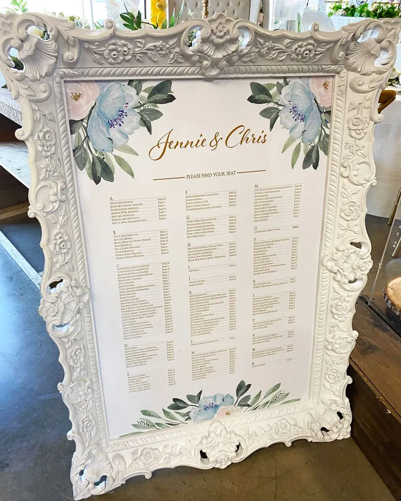 Tableau de plan de table élégant pour le mariage de Jennie et Chris, présenté dans un cadre orné blanc avec des motifs floraux dans les coins, créés par Photogenia Design Studio. Cette invitation de mariage, appartenant à la catégorie des invitations de mariage de Mariage Elegant, présente une liste organisée des places des invités avec des illustrations de fleurs bleues et vertes, offrant un aperçu raffiné et personnalisé pour les célébrations nuptiales au Canada.