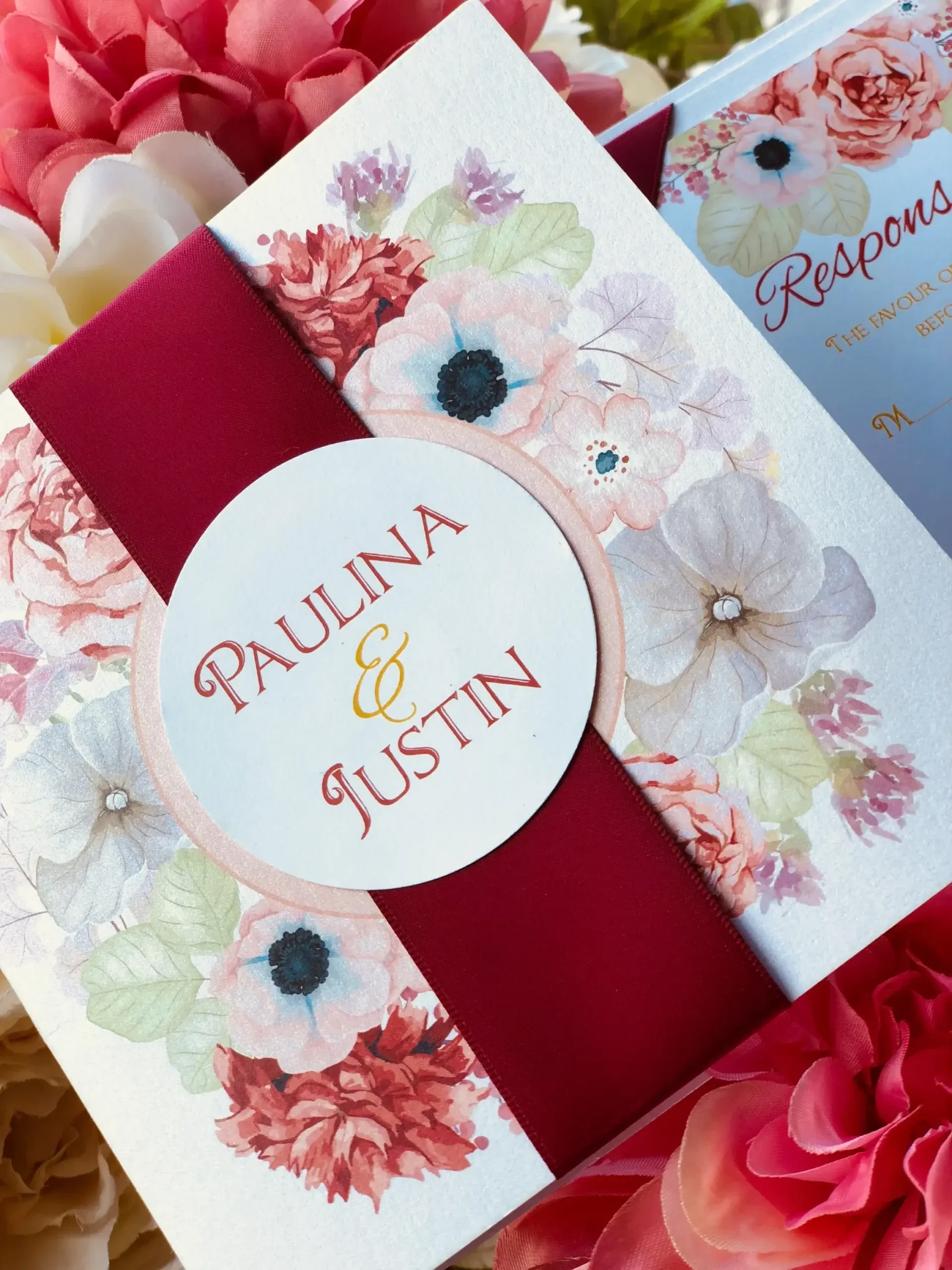 Une élégante invitation de mariage conçue par Photogenia Design Studio, présentée dans la catégorie Invitations de mariage sur Mariage Elegant, Canada. L'invitation est ornée de fleurs colorées dans des tons pastel, comme le rose et le blanc, sur fond crème. Au centre, un bandeau rouge cerise ajoute une touche de couleur profonde et met en valeur les prénoms 'Paulina & Justin' en lettres dorées. L'invitation est posée parmi un arrangement de fleurs roses et blanches, créant un cadre harmonieux et romantique. Ce design est idéal pour un mariage raffiné et élégant.